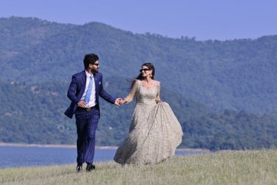 A couple in elegant wedding attire walking hand in hand by a picturesque riverbank in Una, India.